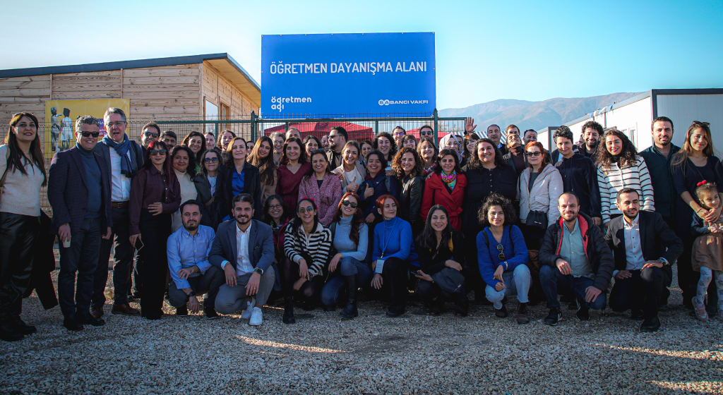 Öğretmen Dayanışma Alanı açılışının sonunda açılış katılımcıları ile çekilen fotoğrafta katılımcılar alanın tabelası altında kameraya poz veriyor. Tabelada “Öğretmen Dayanışma Alanı” yazıyor. Tabelada yazının sol altında Öğretmen Ağı, sağ altında Sabancı Vakfı logosu bulunuyor.